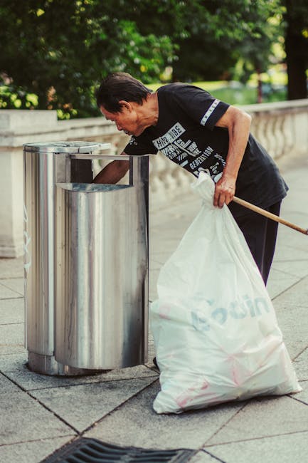 A man with light skin and dark hair, dressed in a black T-shirt with white lettering, is seen leaning over and depositing waste into a cylindrical metal rubbish bin located outdoors on a paved sidewalk. He is holding a large white plastic bag filled with rubbish in his left hand while using his right hand to reach into the bin. The bin is made of stainless steel with a smooth finish and has an open top, positioned close to a low, beige stone or cement wall with a decorative railing behind it. Greenery, including trees and shrubs, can be seen in the background, providing a natural setting for the scene. The lighting is natural, indicating daytime, and the overall scene suggests routine outdoor waste disposal, possibly part of a private or independent rubbish collection effort, aligning with services typically offered by companies such as Rubbish Collection Acton.