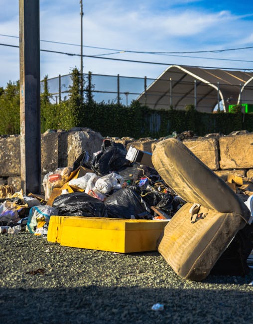 A collection of discarded rubbish piled on the ground beside a low stone wall, including multiple black and grey garbage bags, a yellow plastic container, and an upright dirty, worn shoe. In the background, a chain-link fence surrounds a sports court with a partially visible white canopy structure, and a blue sky with some wispy clouds is overhead. The scene appears to be outdoors on a gravel or asphalt surface, with utility poles and power lines visible, suggesting an urban or suburban environment where private waste disposal such as rubbish removal services may be needed, as indicated by the presence of waste typical of on-site clearance or alternative collection options offered by companies like Rubbish Collection Acton.