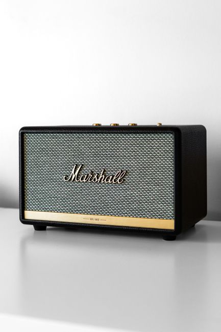 A rectangular Marshall portable speaker with a textured front grille in light grey and a metallic gold logo centered on the front, set against a plain, light-colored surface and a neutral background. The speaker features a black plastic casing with rounded edges, four small rubber feet at the bottom, and control knobs on the top edge. The overall appearance is sleek and modern, with a combination of matte black and metallic finishes. The image highlights the device’s design and build, typical of audio equipment that could be associated with media and entertainment environments, indirectly relating to waste management contexts through the neutral setting and absence of clutter or other objects.