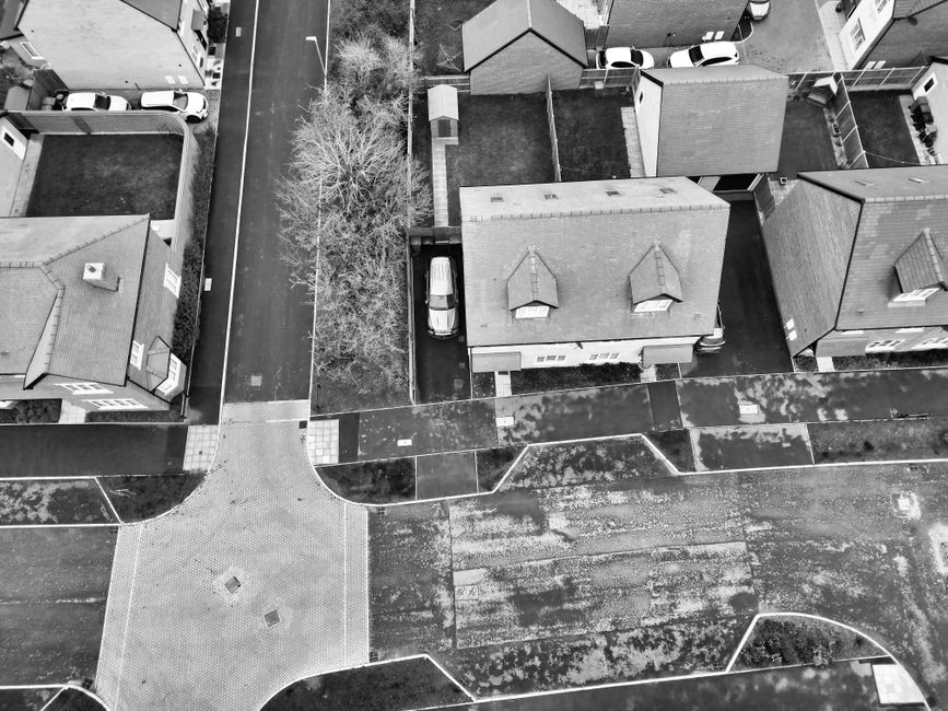 An aerial view of a residential area showing multiple houses with pitched roofs, primarily composed of tiles, surrounded by paved pathways and small gardens. In the center of the image, there is a driveway with a parked van positioned next to a narrow backyard, which contains a leafless tree and a small shed or garage. Surrounding the houses are driveways and parking areas with a few cars visible along the edges of the properties. The pavement and pathways exhibit a combination of smooth concrete and textured brick surfaces, with some patches of moss or dirt visible on the ground. The scene is captured in black and white, emphasizing the textures of the roofing materials, paving, and foliage, indicative of a typical suburban setting. This aerial perspective highlights the layout of the properties and the surrounding infrastructure, relating to private waste management and alternative rubbish collection services like those offered by Rubbish Collection Acton, for effective on-site clearance and waste handling in residential estates.