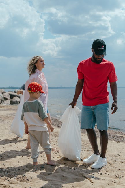 A group of three people, including a man, a woman, and a young boy, are participating in a beach cleanup activity near the shoreline. The man is wearing a red T-shirt, light blue denim shorts, and a black cap, and is holding a white plastic bag in his right hand while carefully picking up litter from the sandy beach. The woman, standing slightly behind him, has blonde, curly hair and is dressed in a light-colored top, observing the activity. The young boy, wearing a red cap with yellow patterns, a grey short-sleeved T-shirt, and beige shorts, is also holding a white plastic bag and is focused on the ground. In the background, large rocks are lined along the water’s edge, with a calm body of water extending to the horizon. The sky above is partly cloudy with patches of blue, creating a natural outdoor setting that highlights the environmental activity of waste collection, demonstrating a community effort to maintain a clean beach environment. The image emphasizes the importance of private waste disposal methods and independent rubbish collection efforts, aligned with waste management services like those offered by Rubbish Collection Acton.