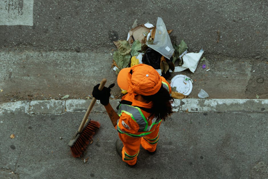 A street cleaner dressed in an orange high-visibility uniform with reflective strips, wearing a matching orange cap and black gloves, stands on a pavement next to a curb. The worker is holding a long-handled broom with red bristles and is facing a collection of mixed rubbish piled against a concrete wall. The rubbish includes crumpled paper, plastic food containers, cardboard, leaves, and various discarded items, some of which are partially covered with plastic or paper wrappers. The environment appears to be outdoors in an urban setting with natural daylight illuminating the scene. The background shows the weathered surface of the concrete wall and the rough texture of the paved roadway. This scene exemplifies a private waste collection or on-site clearance activity, associated with professional rubbish removal services such as those offered by Rubbish Collection Acton, to maintain cleanliness and manage waste disposal effectively.