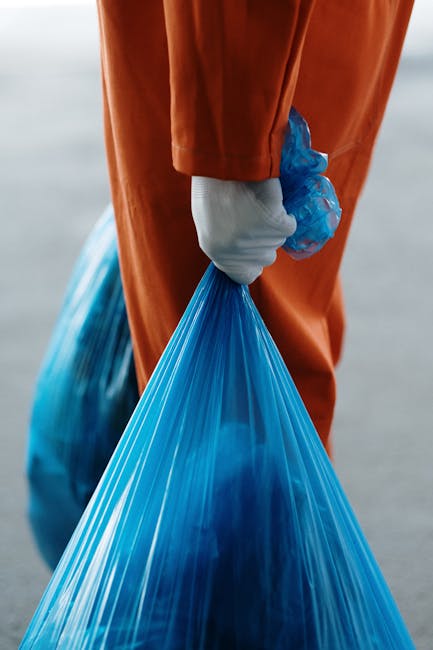 A group of three people, including a man, a woman, and a young boy, are participating in a beach cleanup activity near the shoreline. The man is wearing a red T-shirt, light blue denim shorts, and a black cap, and is holding a white plastic bag in his right hand while carefully picking up litter from the sandy beach. The woman, standing slightly behind him, has blonde, curly hair and is dressed in a light-colored top, observing the activity. The young boy, wearing a red cap with yellow patterns, a grey short-sleeved T-shirt, and beige shorts, is also holding a white plastic bag and is focused on the ground. In the background, large rocks are lined along the water’s edge, with a calm body of water extending to the horizon. The sky above is partly cloudy with patches of blue, creating a natural outdoor setting that highlights the environmental activity of waste collection, demonstrating a community effort to maintain a clean beach environment. The image emphasizes the importance of private waste disposal methods and independent rubbish collection efforts, aligned with waste management services like those offered by Rubbish Collection Acton.